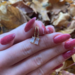 A close-up of a person's hand with red nail polish, wearing a pair of gold earrings with clear gemstones.