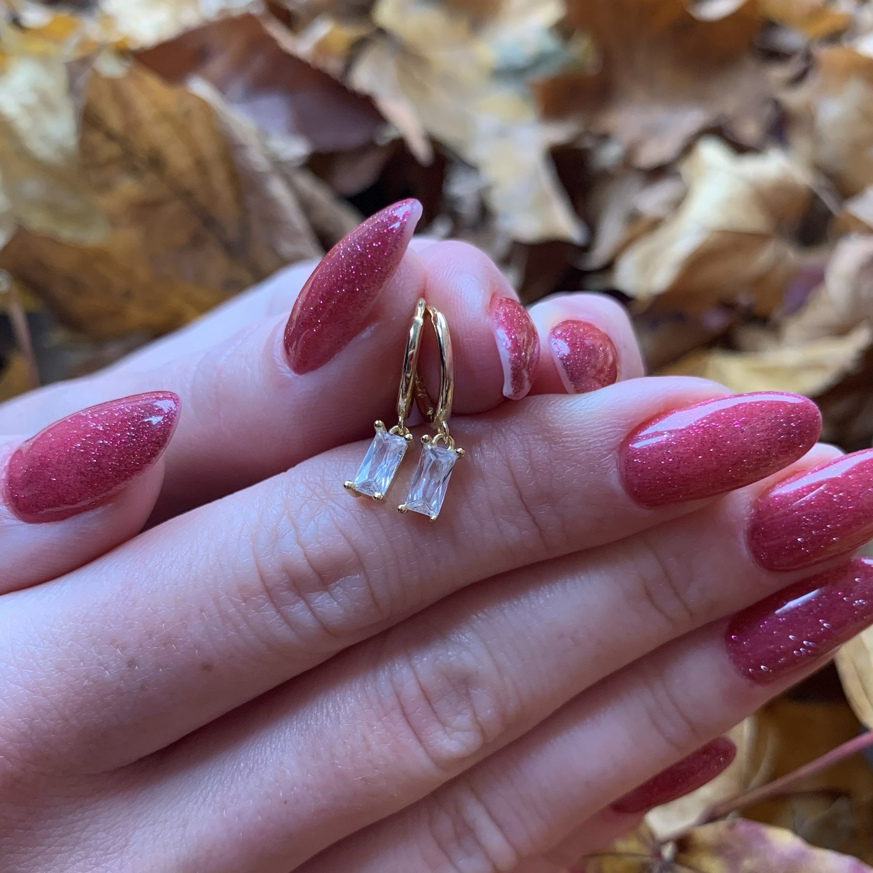 A close-up of a person's hand with red nail polish, wearing a pair of gold earrings with clear gemstones.