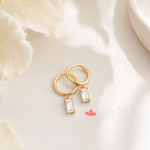 A pair of gold hoop earrings with clear gemstone pendants, resting on a white surface with a floral pattern.