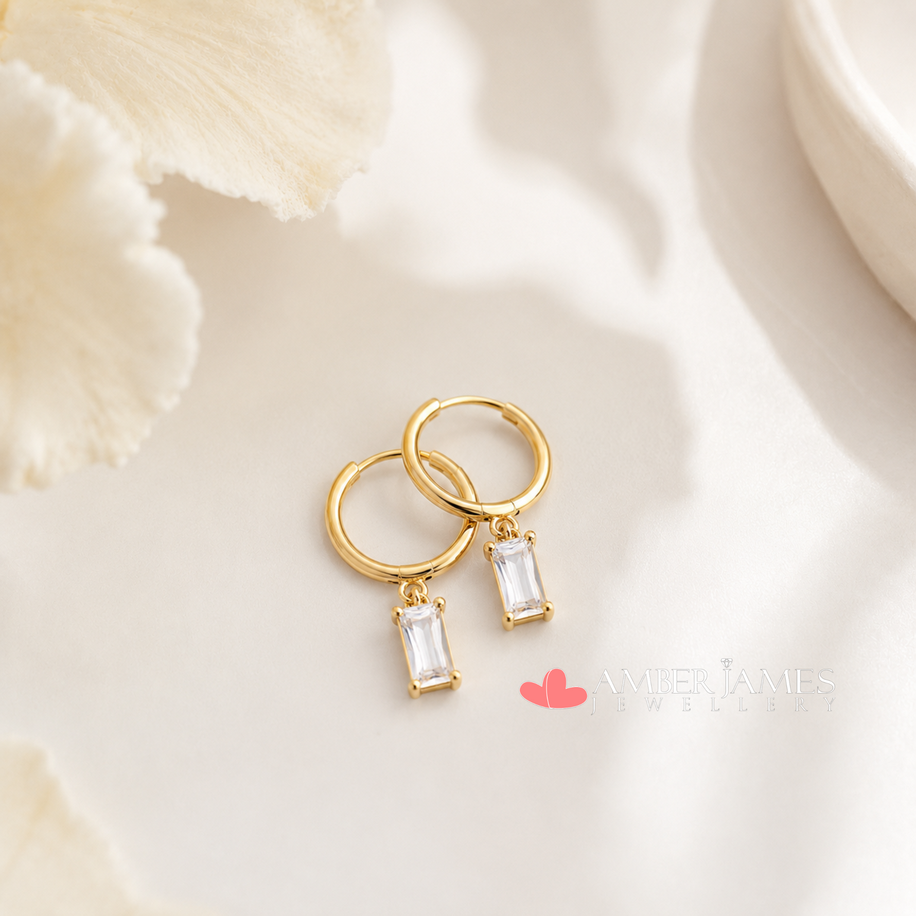 A pair of gold hoop earrings with clear gemstone pendants, resting on a white surface with a floral pattern.