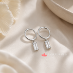 A pair of silver hoop earrings with clear gemstone pendants, resting on a beige fabric surface.