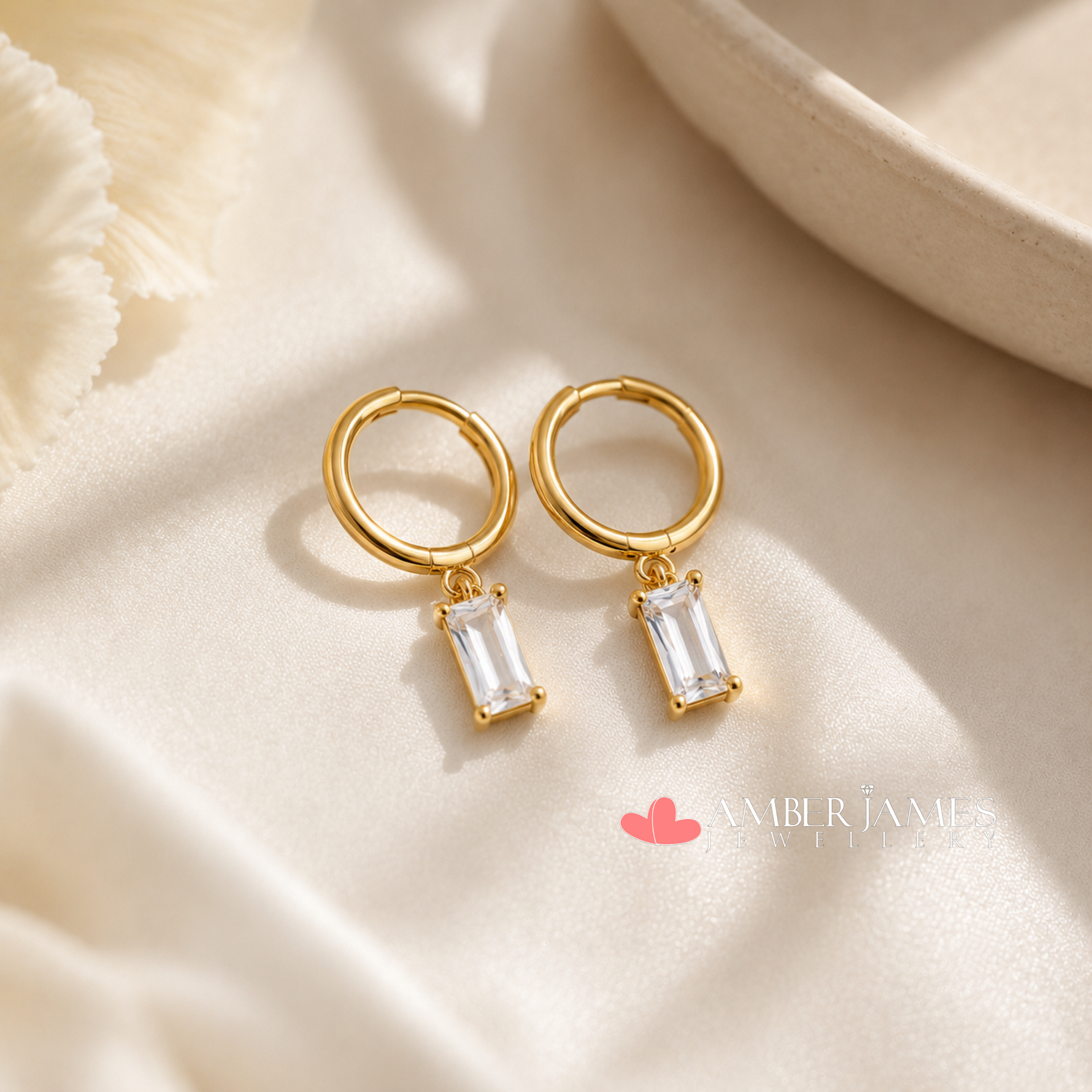 A pair of gold hoop earrings with clear gemstone pendants, placed on a white fabric surface.